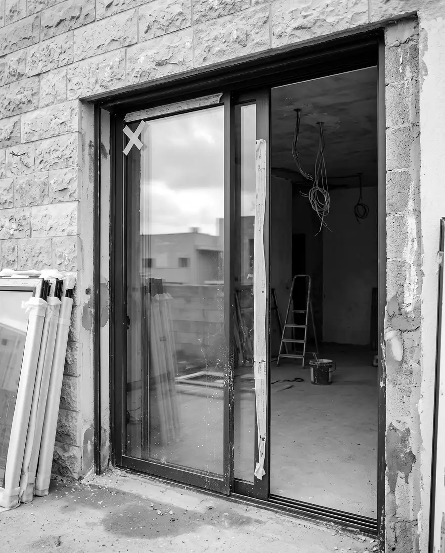 דלת הזזה מאלומיניום שהותקנה בקיר אבן של בית בבנייה: סרט הדבקה על הזכוכית, חוטי חשמל תלויים בפנים, חלונות עטופים בקצף בצד